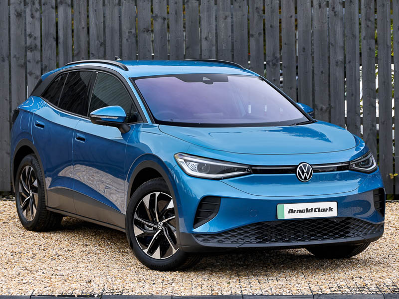 A blue VW ID.4 parked side on, sitting on gravel with a grey fence in the background
